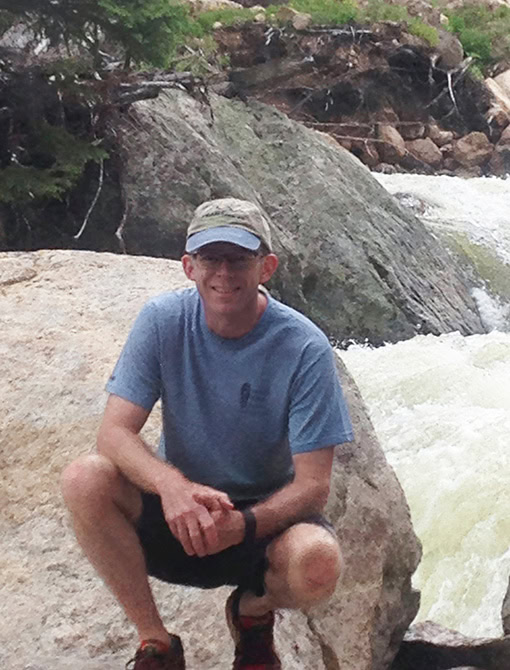 A man in a blue t-shirt and cap crouching on a large rock near a flowing river with rocks and trees in the background, outdoors, during daytime.