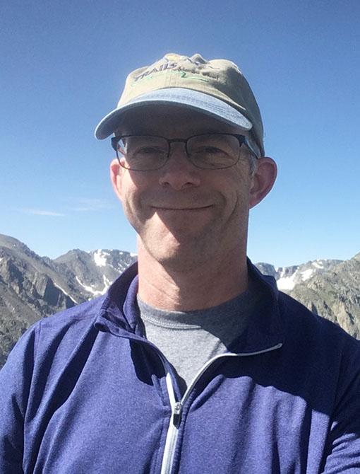 A smiling man wearing glasses, a baseball cap, and a blue jacket outdoors with mountains and a clear blue sky in the background, representing Creve Coeur Behavioral Counseling.