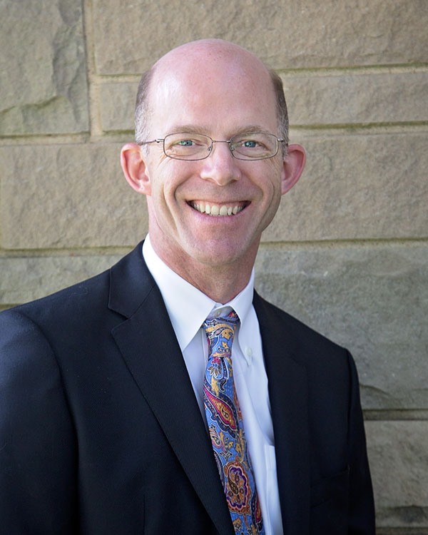 A smiling professional man in a suit and colorful tie standing outdoors against a stone wall, representing the Creve Coeur Behavioral Counseling team, part of the Our Team section.